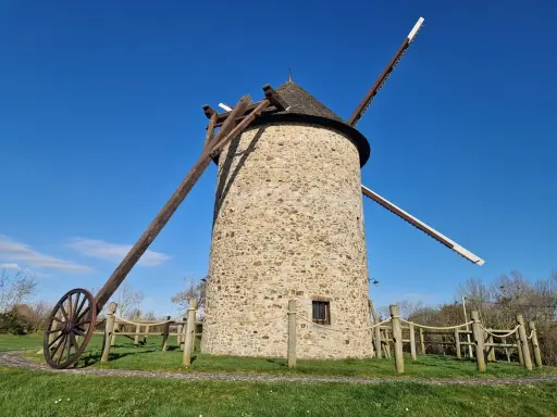 Le moulin de Maryline, une tour de pierre imposante sous un ciel bleu, se dresse fièrement avec ses ailes en bois.