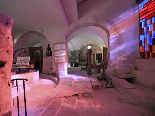 Intérieur d'un musée du verre et du vitrail, avec des arches en pierre, des outils anciens et un vitrail coloré projetant des rayons lumineux.