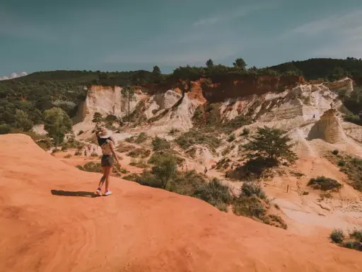 Une femme au chapeau admire le paysage spectaculaire de la carrière d'ocre du Vaucluse, avec ses falaises colorées et sa végétation luxuriante.