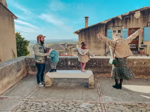 Famille profitant d'une vue panoramique depuis un balcon à Châteauneuf de Gadagne. Le père tient un enfant, tandis que deux autres enfants jouent sur un banc.