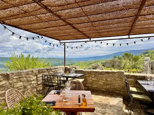 Terrasse d'un restaurant avec vue panoramique sur les collines. Tables dressées sous un auvent de chaume, ambiance estivale. Un jardin sur le toit Saignon promet un repas inoubliable.