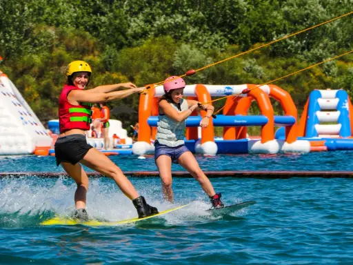 Deux femmes font du wakeboard dans un wam park orange et bleu, avec des structures gonflables en arrière-plan.