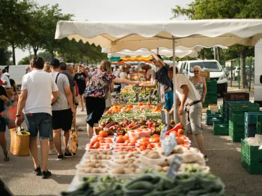 Un marché animé, les angles des étals débordent de fruits et légumes frais. Des clients font leurs courses sous les parasols blancs.