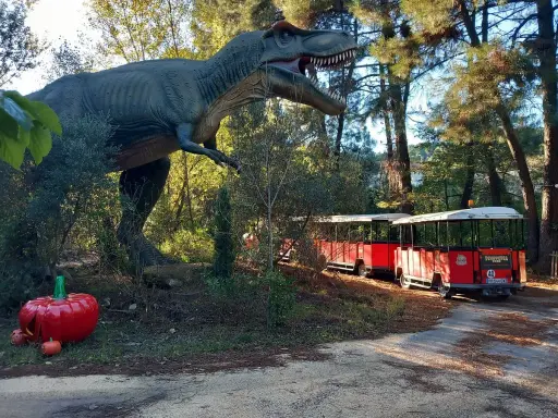 Un train rouge traverse le parc Grimmland, passant près d'un dinosaure géant et de citrouilles décoratives.
