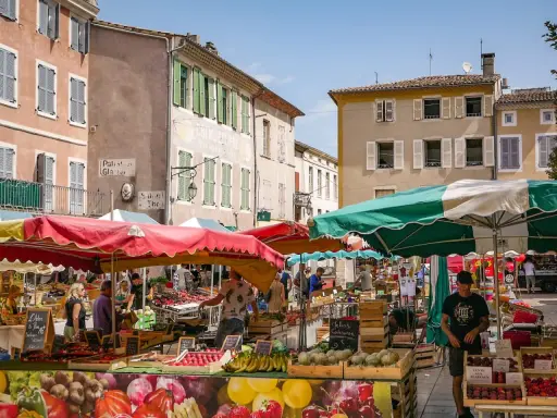 Le marché le Thor animé, avec des étals colorés de fruits et légumes sous des parasols. Des passants flânent entre les stands, profitant du soleil provençal.
