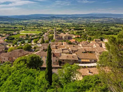 Vue aérienne du village de Bonnieux, avec ses toits de tuiles ocres, son église et la campagne environnante verdoyante. Le marché de Bonnieux est un lieu animé.