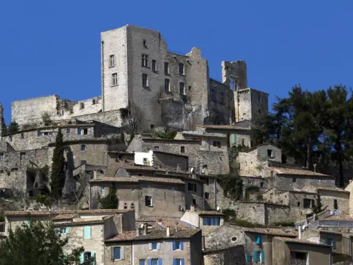 Vue d'un village perché dominé par les ruines imposantes du **château du marquis de Sade**, sous un ciel bleu éclatant.