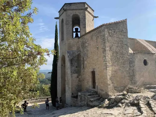 Le clocher de Notre-Dame d'Alidon, avec sa cloche visible, se dresse fièrement sous un ciel bleu. Des visiteurs admirent le paysage.