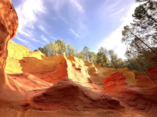 Carrières d'ocre aux couleurs vives, rappelant l'usine d'ocre Mathieu. Le ciel bleu et les arbres verts contrastent avec les roches rouges et jaunes.