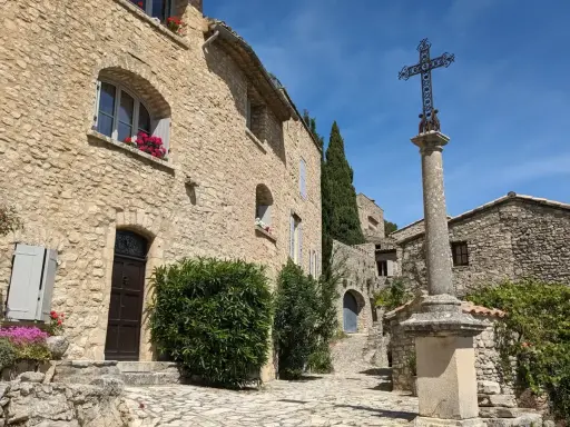 Village de pierre au soleil, le Crestet Vaucluse. Une croix orne la place pavée, entourée de maisons fleuries.