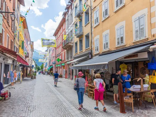 Rue commerçante animée à Aubenas-les-Alpes, avec des façades colorées, des drapeaux et des passants profitant de l'été.