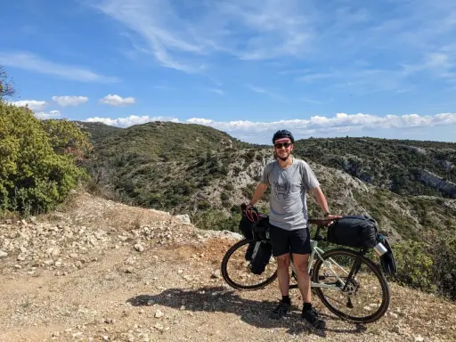 Cycliste souriant avec son vélo chargé, profitant d'une pause lors d'un tour du Luberon à vélo. Paysage rocailleux et collines verdoyantes sous un ciel bleu.