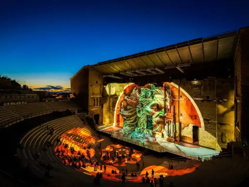 Spectacle son et lumière dans un théâtre antique. Le parvis orange illumine les gradins où le public est assis, tandis que la scène projette une fresque monumentale.