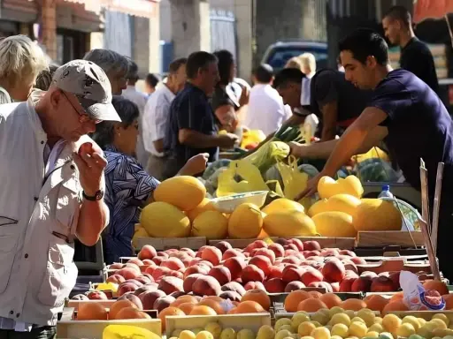 Ambiance animée au marché de Pernes-les-Fontaines. Des étals débordent de fruits colorés, pêches, melons, et agrumes. Des clients choisissent leurs produits.