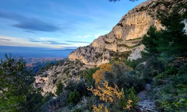 Vue panoramique des gorges de Badarel, avec un sentier rocailleux serpentant à travers la végétation méditerranéenne et une falaise imposante baignée de soleil.