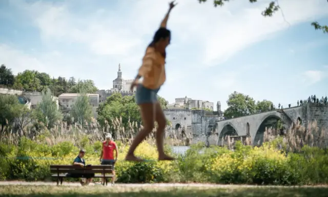 Amusez-vous à Avignon ce week-end ! Une jeune femme saute devant le Palais des Papes et le Pont d'Avignon.