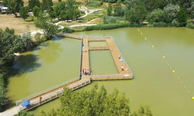 Ponton en bois sur un lac verdâtre, une **base de loisirs** animée avec des baigneurs profitant du soleil.