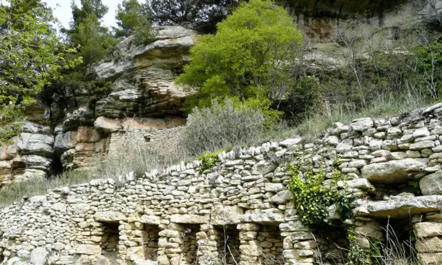 Vestiges d'une ancienne ville de murs en pierre, intégrée à une falaise rocheuse, avec végétation luxuriante.