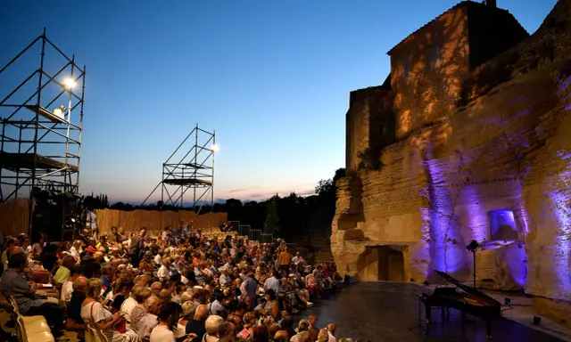 Public venu pour le festival de Gordes, une foule assise attend un concert au crépuscule, avec un piano sur scène et des structures métalliques.