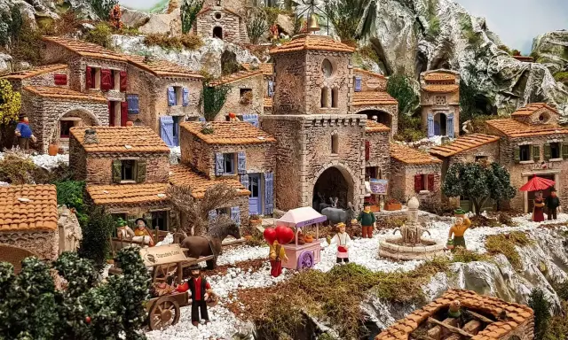 Une **crèche provençale** animée avec des santons, des maisons en pierre, un âne, un chariot et une fontaine, le tout dans un paysage rocheux.