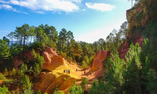 Des randonneurs explorent le sentier des ocres, un paysage spectaculaire aux falaises rouges et jaunes, bordé de pins verts sous un ciel bleu.