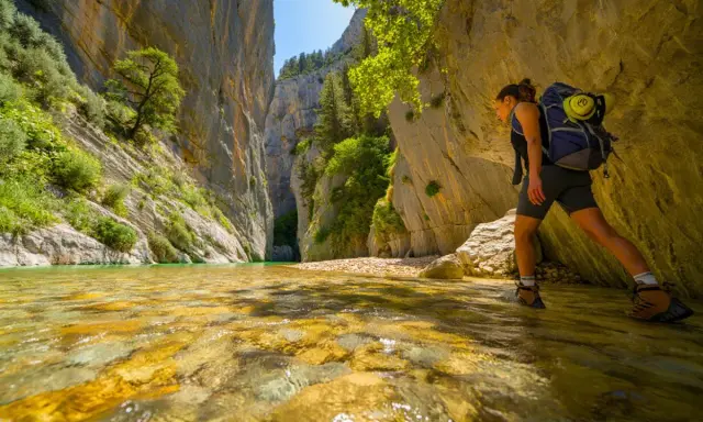 Randonneuse explorant la gorge du Toulourenc, traversant une rivière aux eaux cristallines bordée de falaises imposantes et de végétation luxuriante.