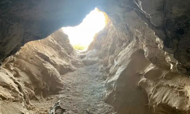 L'entrée de la grotte de Saint Eucher s'ouvre sur une lumière vive, révélant des parois rocheuses sculptées par le temps. Un morceau de bois mort repose au sol.