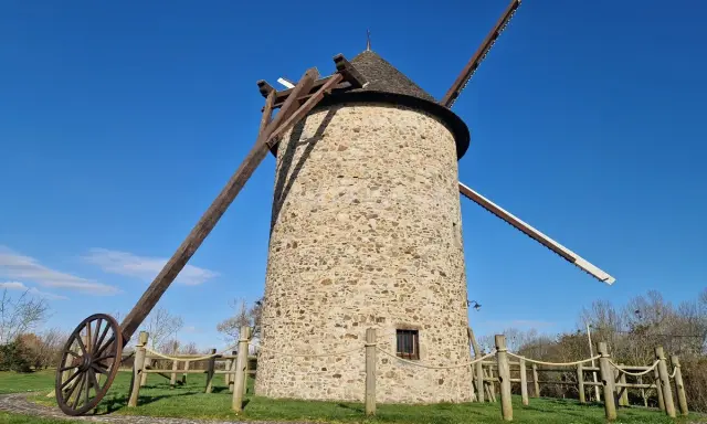 Le moulin de Maryline, une tour de pierre imposante sous un ciel bleu, se dresse fièrement avec ses ailes en bois.