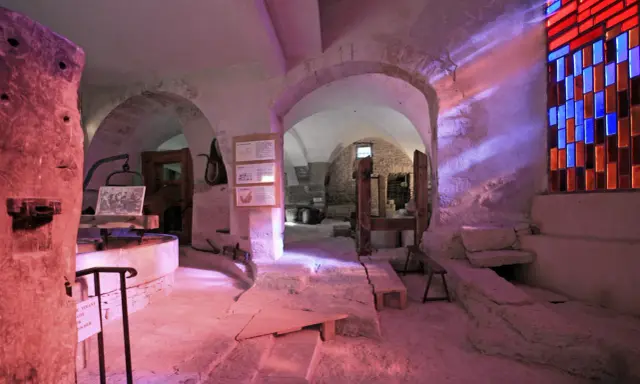 Intérieur d'un musée du verre et du vitrail, avec des arches en pierre, des outils anciens et un vitrail coloré projetant des rayons lumineux.