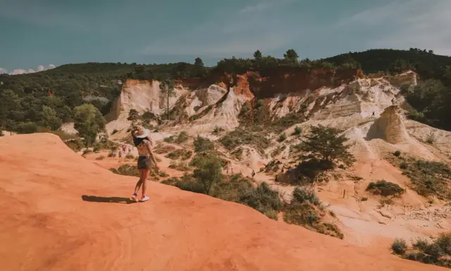 Une femme au chapeau admire le paysage spectaculaire de la carrière d'ocre du Vaucluse, avec ses falaises colorées et sa végétation luxuriante.
