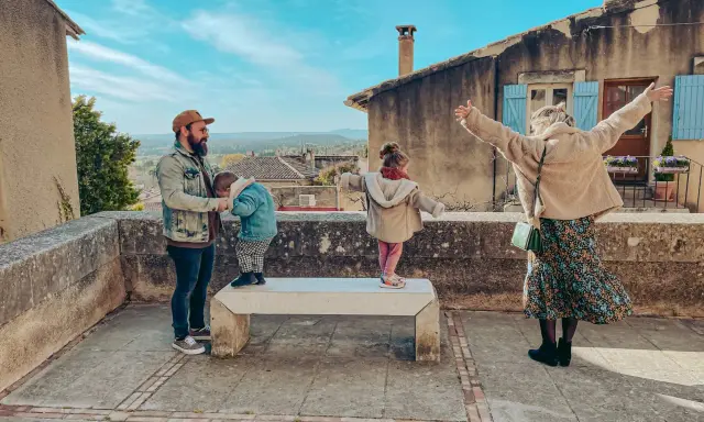 Famille profitant d'une vue panoramique depuis un balcon à Châteauneuf de Gadagne. Le père tient un enfant, tandis que deux autres enfants jouent sur un banc.