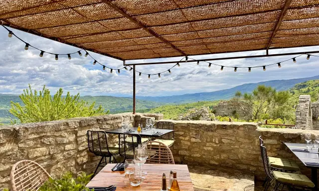 Terrasse d'un restaurant avec vue panoramique sur les collines. Tables dressées sous un auvent de chaume, ambiance estivale. Un jardin sur le toit Saignon promet un repas inoubliable.