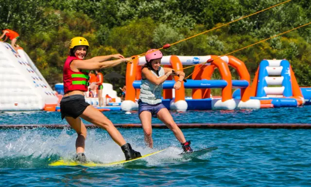 Deux femmes font du wakeboard dans un wam park orange et bleu, avec des structures gonflables en arrière-plan.