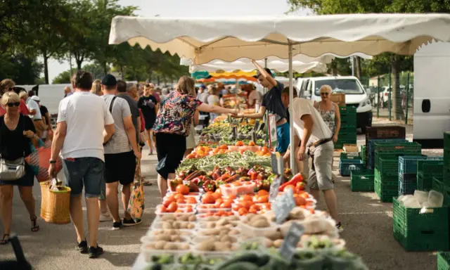 Un marché animé, les angles des étals débordent de fruits et légumes frais. Des clients font leurs courses sous les parasols blancs.