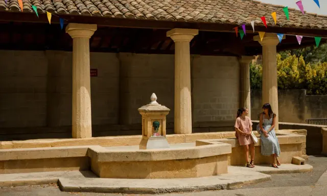 Deux femmes discutent près d'une fontaine sous un auvent orné de fanions colorés, ambiance de marché à Rochefort-du-Gard.