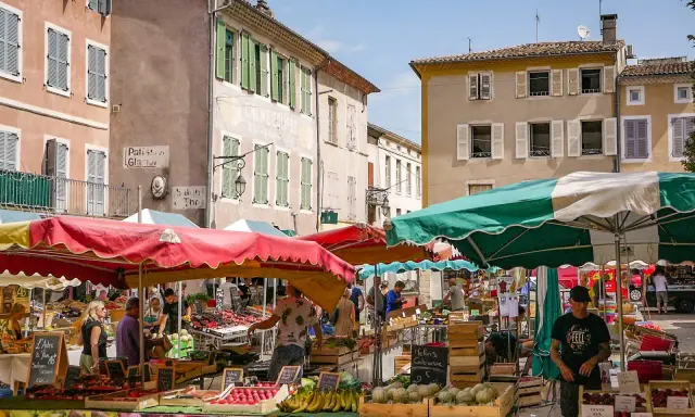 Le marché le Thor animé, avec des étals colorés de fruits et légumes sous des parasols. Des passants flânent entre les stands, profitant du soleil provençal.