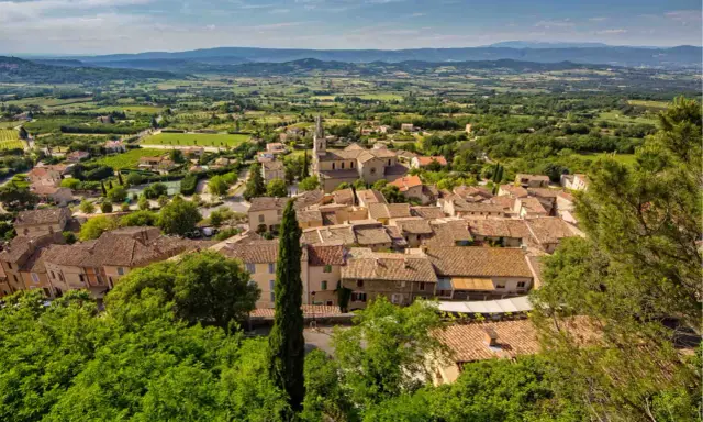 Vue aérienne du village de Bonnieux, avec ses toits de tuiles ocres, son église et la campagne environnante verdoyante. Le marché de Bonnieux est un lieu animé.