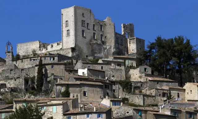 Vue d'un village perché dominé par les ruines imposantes du **château du marquis de Sade**, sous un ciel bleu éclatant.