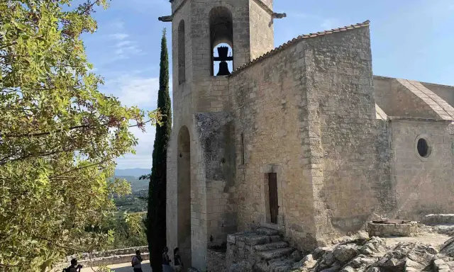 Le clocher de Notre-Dame d'Alidon, avec sa cloche visible, se dresse fièrement sous un ciel bleu. Des visiteurs admirent le paysage.