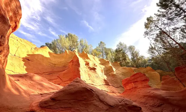 Carrières d'ocre aux couleurs vives, rappelant l'usine d'ocre Mathieu. Le ciel bleu et les arbres verts contrastent avec les roches rouges et jaunes.