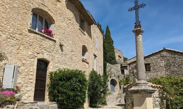Village de pierre au soleil, le Crestet Vaucluse. Une croix orne la place pavée, entourée de maisons fleuries.