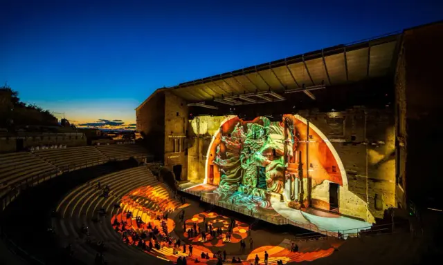 Spectacle son et lumière dans un théâtre antique. Le parvis orange illumine les gradins où le public est assis, tandis que la scène projette une fresque monumentale.