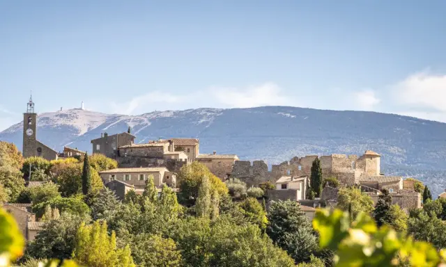 Vue d'un faucon village perché sur une colline, avec une église et des ruines médiévales. Le Mont Ventoux domine l'horizon.