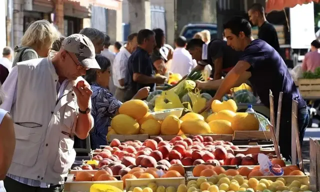 Ambiance animée au marché de Pernes-les-Fontaines. Des étals débordent de fruits colorés, pêches, melons, et agrumes. Des clients choisissent leurs produits.