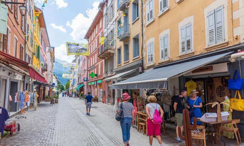 Rue commerçante animée à Aubenas-les-Alpes, avec des façades colorées, des drapeaux et des passants profitant de l'été.