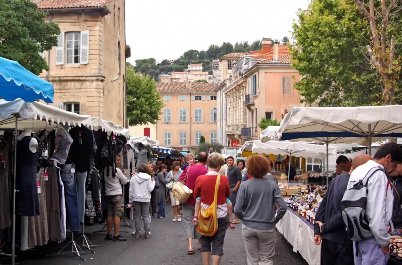 Ambiance animée au marché d'Apt, avec des étals de vêtements et de produits locaux. Des passants flânent sous les parasols, profitant de l'atmosphère provençale.