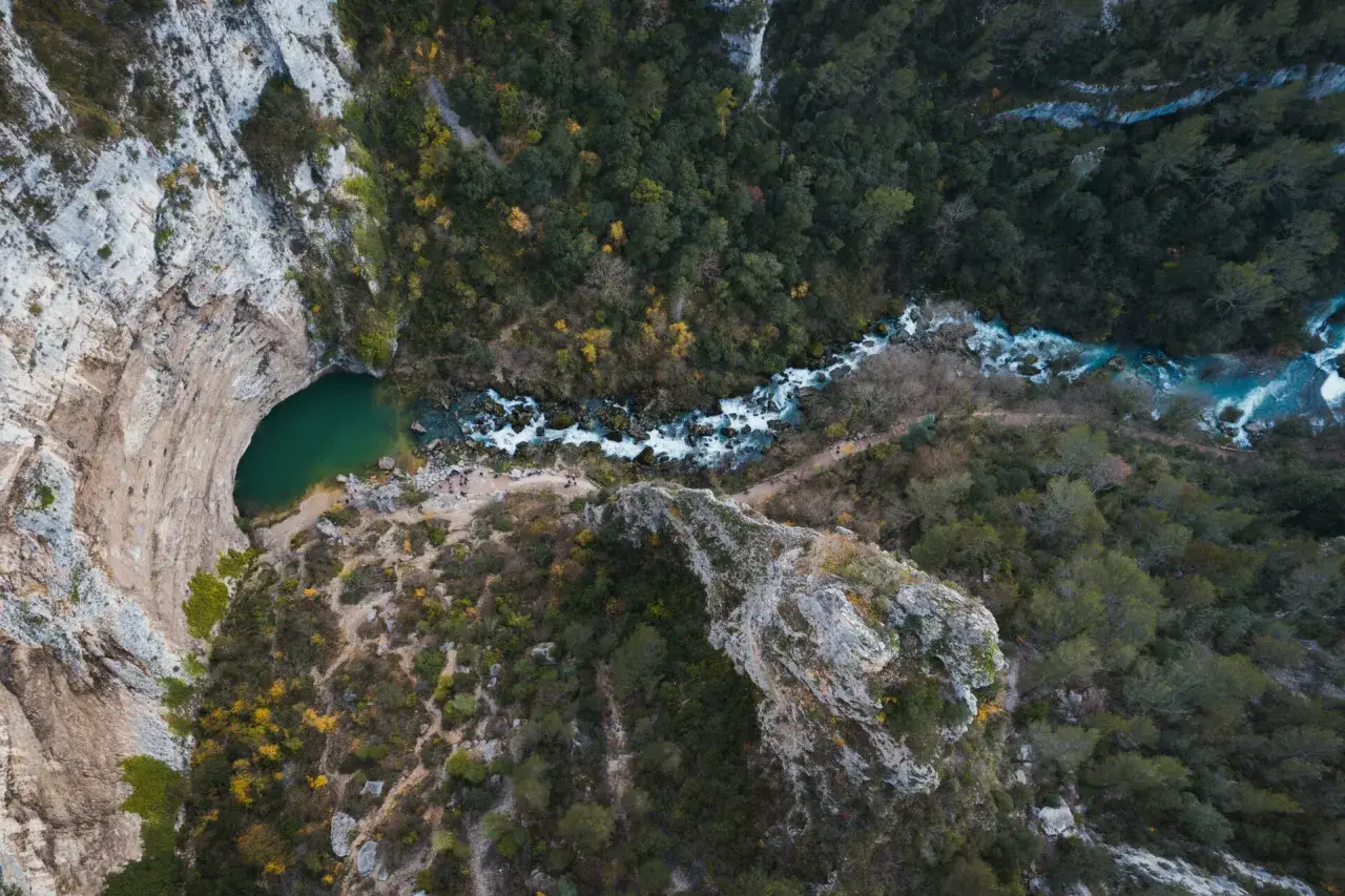 Vue aérienne de la Fontaine de Vaucluse, avec son bassin émeraude et la rivière Sorgue serpentant à travers une gorge boisée.