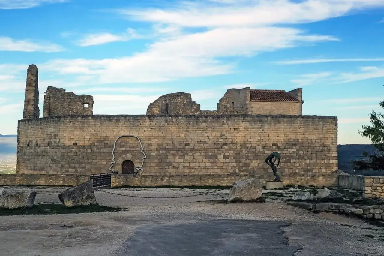 Le ch&acirc;teau du marquis de Sade, en ruines, se dresse sous un ciel bleu. Une sculpture de t&ecirc;te humaine orne le mur de pierre.