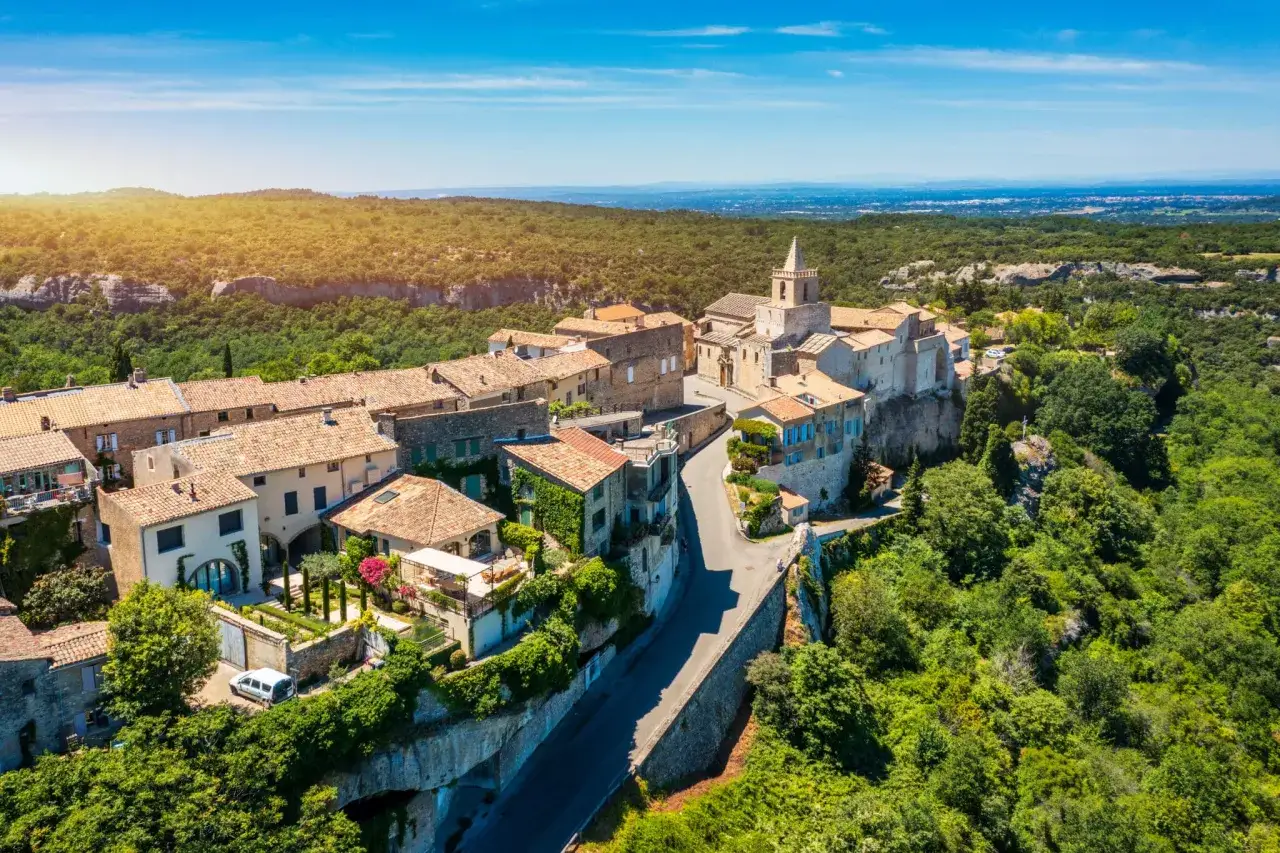 Vue a&eacute;rienne du village perch&eacute; de Venasque, Vaucluse, avec ses maisons aux toits de tuiles, son &eacute;glise et la verdure environnante sous un ciel bleu.