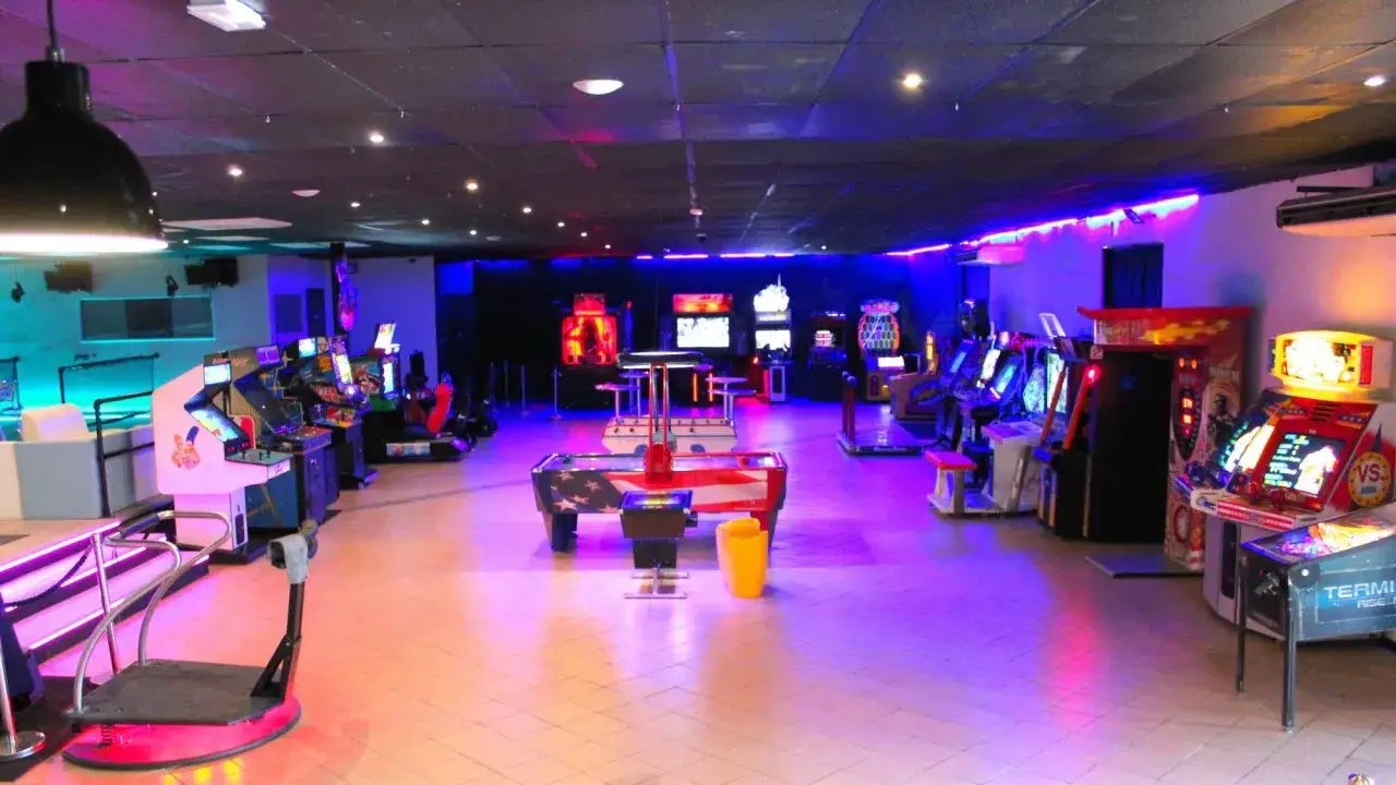 Salle de jeux vid&eacute;o illumin&eacute;e de n&eacute;ons bleus et roses, avec des bornes d'arcade et un baby-foot. L'ambiance rappelle l'elyzion orange des soir&eacute;es d'&eacute;t&eacute;.